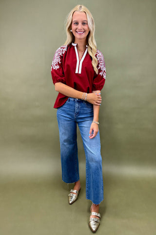 Woman wearing a red blouse with white embroidery and blue jeans against a green background