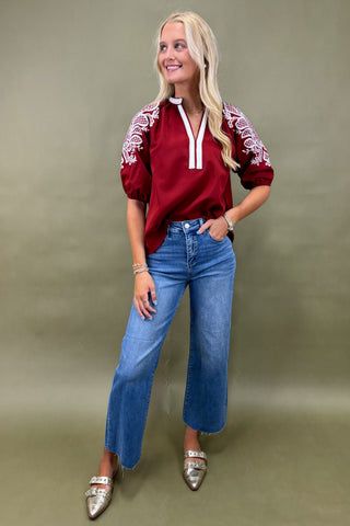 Woman wearing a red blouse with white embroidery and blue jeans against a green background