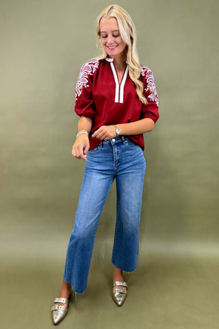 Woman wearing a red blouse with white embroidery and blue jeans against a green background