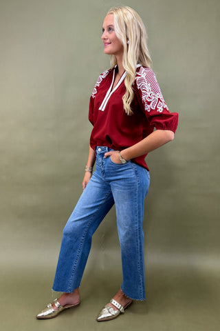 Woman wearing a red blouse with white embroidery and blue jeans against a green background