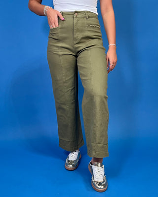 A person wearing olive green wide-leg cropped pants with decorative zigzag stitching on the front, paired with a white top and metallic sneakers
