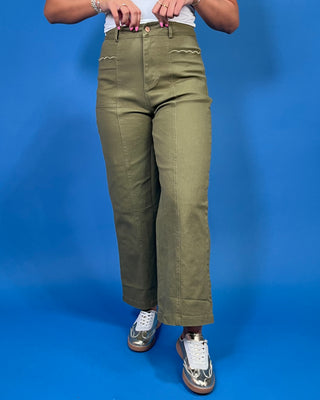 A person wearing olive green wide-leg cropped pants with decorative zigzag stitching on the front pockets, paired with a white top and metallic sneakers