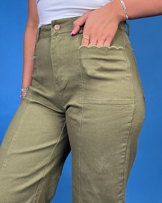 A close-up of a person wearing olive green high-waisted pants with a scalloped edge detail around the pocket, paired with a white top