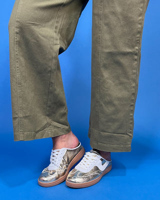 A person wearing olive green wide-leg pants and metallic gold and white slip-on sneakers