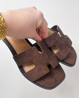 Brown sandal with a hand holding it on a light gray background
