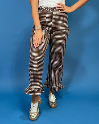 A person modeling a pair of brown and white houndstooth patterned cropped pants with ruffled hems, paired with a white top and metallic silver sneakers