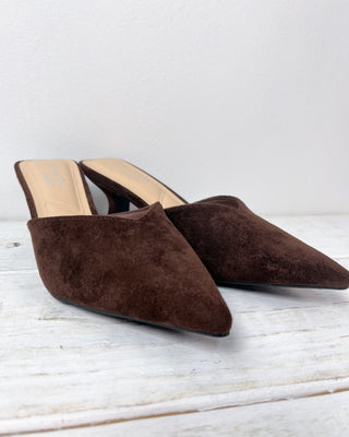 Pair of brown suede mules on a light wooden surface with a white background