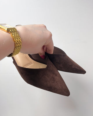Brown pointed-toe shoes held by a hand with gold bracelet on a white background