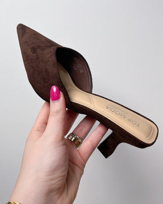 Brown pointed-toe shoe held by a hand with pink nail polish on a light gray background
