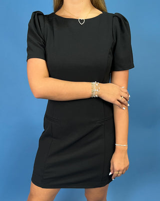 A woman wearing a black short-sleeve dress with puff sleeves and a small silver heart pendant necklace, standing against a solid blue background.