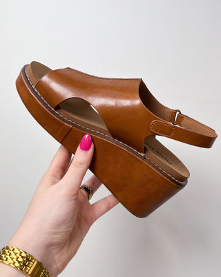 Brown sandal held by a hand with pink nail polish against a light background