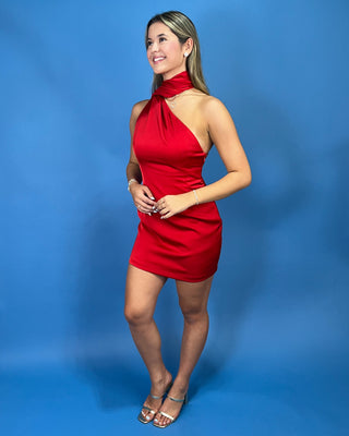 A woman modeling a bright red halter-neck mini dress with a cutout back, standing against a solid blue studio backdrop.