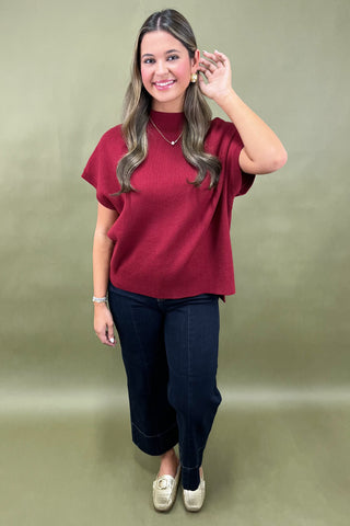 Woman wearing a red top and dark pants against a plain background