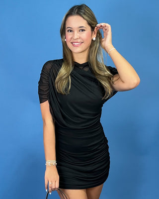 A woman modeling a black ruched mini dress with sheer short sleeves, standing against a solid blue background.