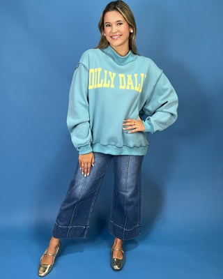 A woman modeling a light blue sweatshirt with yellow 'DILLY DALLY' text, paired with wide-leg denim jeans and metallic flats, against a solid blue studio backdrop.