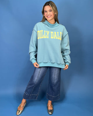 A woman modeling a light blue sweatshirt with yellow 'DILLY DALLY' text, paired with wide-leg denim jeans and metallic flats, against a solid blue studio backdrop.