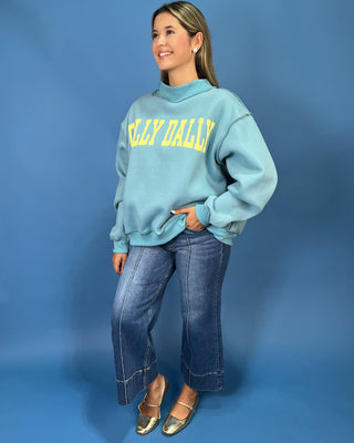 A woman modeling a light blue sweatshirt with yellow 'DILLY DALLY' text, paired with wide-leg denim jeans and metallic flats, against a solid blue studio backdrop.