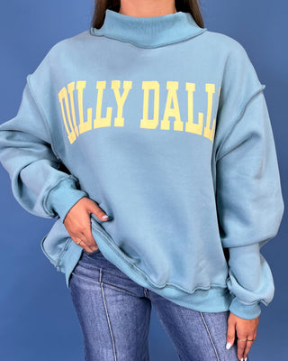 A woman modeling a light blue sweatshirt with yellow 'DILLY DALLY' text, paired with wide-leg denim jeans and metallic flats, against a solid blue studio backdrop.