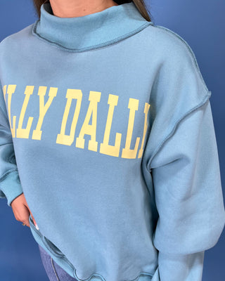 A woman modeling a light blue sweatshirt with yellow 'DILLY DALLY' text, paired with wide-leg denim jeans and metallic flats, against a solid blue studio backdrop.