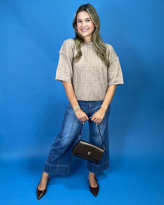 A woman modeling a textured beige short-sleeve knit top, blue wide-leg cropped jeans, and black pointed-toe heels, holding a black leather handbag against a solid blue studio backdrop.