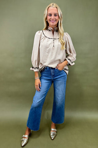 Woman wearing a beige blouse with floral embroidery and blue jeans against a green background