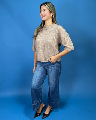 A woman modeling a textured beige short-sleeve knit top and blue wide-leg jeans against a solid blue studio background.