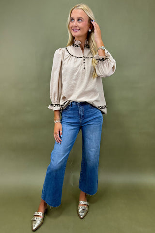 Woman wearing a beige blouse with floral embroidery and blue jeans against a green background