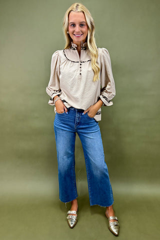 Woman wearing a beige blouse with floral embroidery and blue jeans against a green background