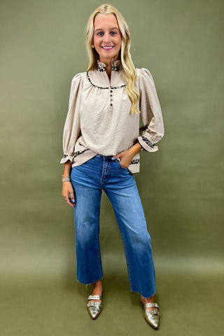 Woman wearing a beige blouse with floral embroidery and blue jeans against a green background