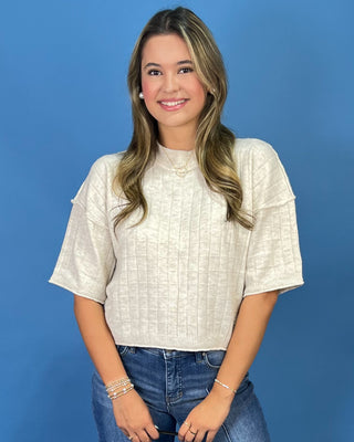 A woman with long blonde-highlighted hair wearing a textured cream short-sleeve crop top and blue jeans, standing against a solid blue studio background.