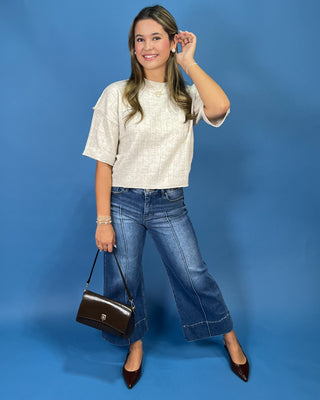 A woman posing in a cream textured short-sleeve top, blue wide-leg jeans, and dark brown pointed-toe flats, holding a dark brown leather handbag against a solid blue studio background.