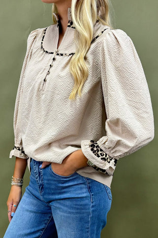 Woman wearing a beige textured blouse with leopard print cuffs and trim, paired with blue jeans against a green background.