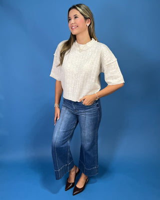 A woman modeling a short-sleeve textured cream linen top and wide-leg blue jeans against a solid blue studio backdrop.