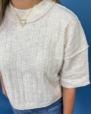 A close-up of a person wearing a cream-colored textured knit mock neck top with short sleeves, paired with blue jeans and a gold necklace, set against a solid blue background.