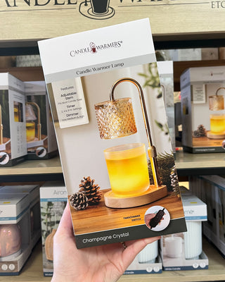 A hand holds a package of Candle Warmers Champagne Crystal candle warmer lamp in a retail store setting with shelves of other products in the background.