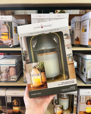 A hand holds a Candle Warmers Golden Glow candle warmer lantern and candle gift set box in a retail store setting with shelves of other products in the background.