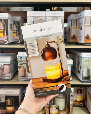 A hand holds a Candle Warmer Lamps 'Brittle Sphere' retail box in front of a store shelf filled with other boxed home decor products.