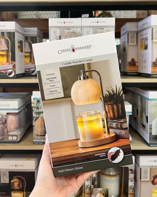 A hand holds a Candle Warmers candle warmer lamp box in a retail store setting. The packaging shows a swan-shaped blown glass lamp with a warm glowing candle inside, set against a background of store shelves filled with other products.