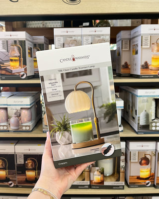 A hand holds a package of Candle Warmers Milk Classa Candle Vase Lamp in front of a store shelf filled with other boxed home decor products.