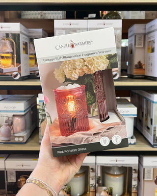 A hand holds a package of Candle Warmers 'Pink Parisian Glass' fragrance warmer in a retail store setting with other products on the shelves behind it.