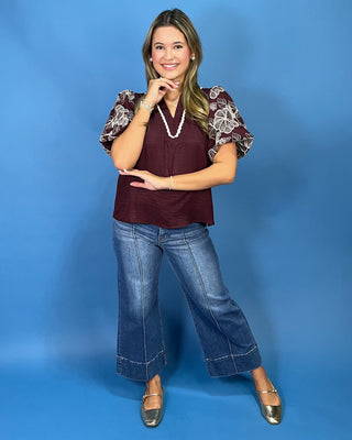 A woman modeling a burgundy short-sleeve blouse with floral lace puff sleeves, paired with blue jeans and metallic flats, standing against a solid blue studio backdrop.