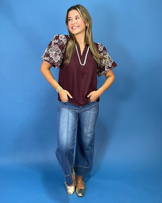 A woman modeling a burgundy short-sleeve top with white floral lace puff sleeves, paired with blue jeans and gold flats, standing against a solid blue studio backdrop.
