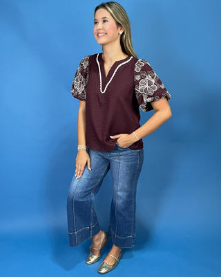 A woman modeling a burgundy short-sleeve top with white floral embroidery on the sleeves and a white beaded V-neck trim, paired with blue jeans and metallic gold flats, against a solid blue studio background.