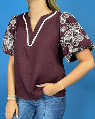 A woman wearing a maroon short-sleeve blouse with white floral puff sleeves and a white scalloped V-neck trim, paired with blue jeans against a solid blue background.