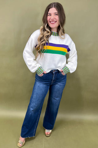 Woman wearing a white sweater with colorful stripes and blue jeans against a beige background