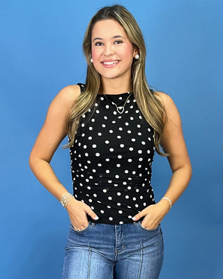 A woman modeling a sleeveless black top with white polka dots, paired with blue jeans, standing against a solid blue background.