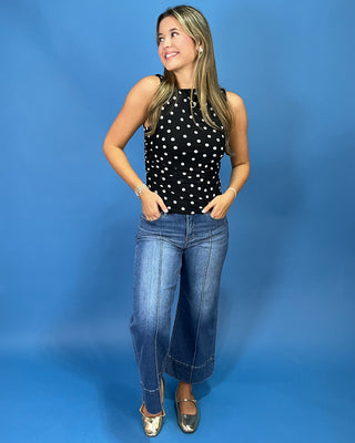 A woman modeling a sleeveless black top with white polka dots, paired with wide-leg blue jeans and metallic gold flats, standing against a solid blue studio backdrop.
