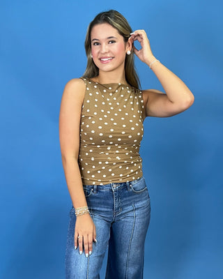 A woman modeling a sleeveless brown top with white polka dots, paired with blue jeans, standing against a solid blue studio background.