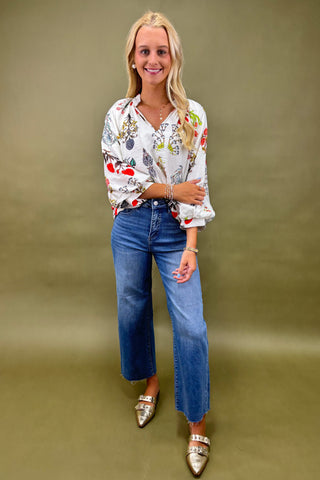 Woman wearing a patterned blouse, blue jeans, and silver shoes against a green background