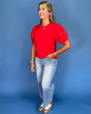 Woman wearing a red blouse with lace sleeves and light blue jeans against a blue background